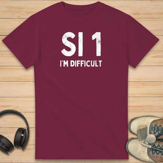 SI 1 T-Shirt in Maroon on a light wooden floor lying next to headphones and golf shoes.