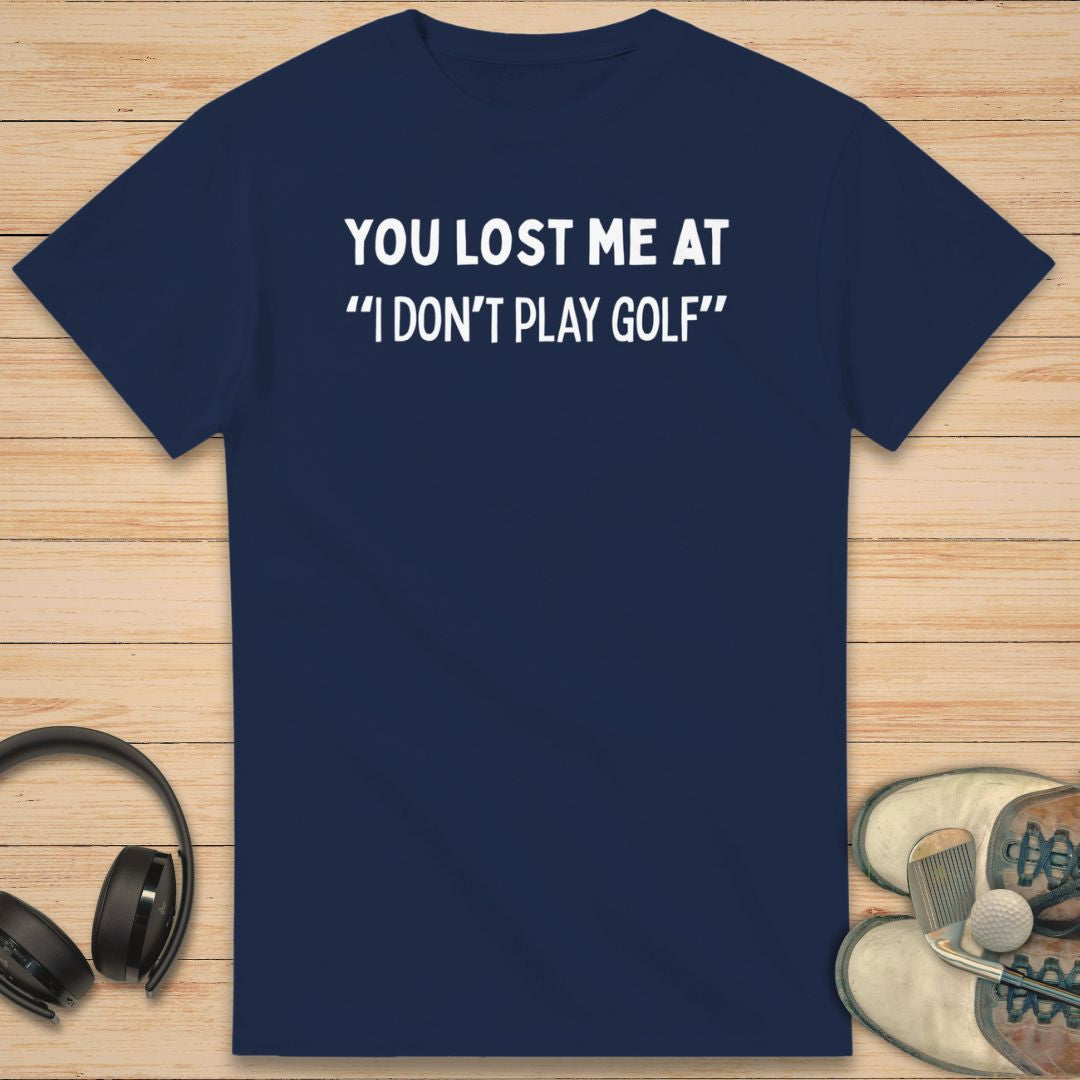 Lost Me T-Shirt in Navy on a light wooden floor lying next to headphones and golf shoes.