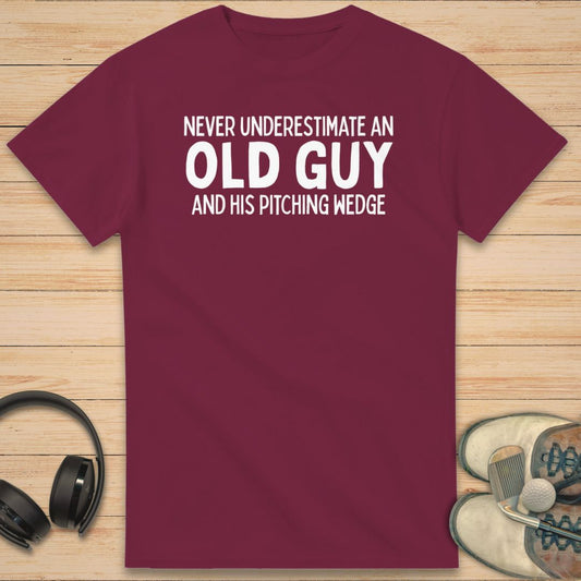 Old Guy T-Shirt in Maroon on a light wooden floor lying next to headphones and golf shoes.