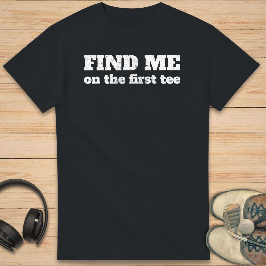 Find Me T-Shirt in Black on a light wooden floor lying next to headphones and golf shoes.