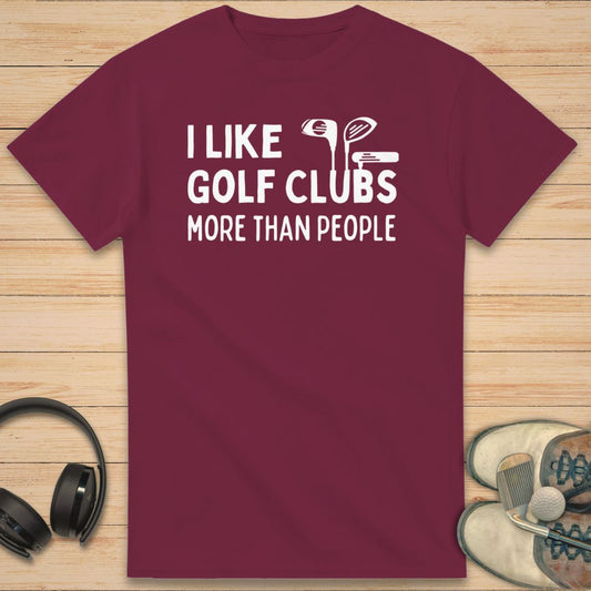 Like Golf Clubs T-Shirt in Maroon on a light wooden floor lying next to headphones and golf shoes.