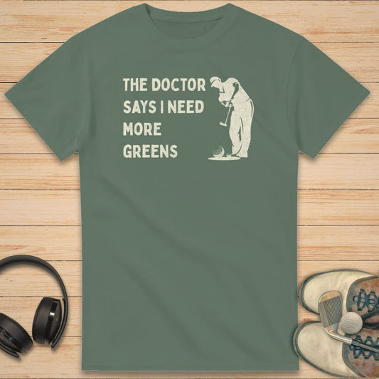 More Greens T-Shirt in Military Green on a light wooden floor lying next to headphones and golf shoes.