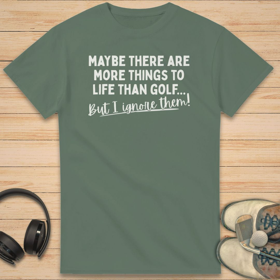 More Things T-Shirt in Military Green on a light wooden floor lying next to headphones and golf shoes.