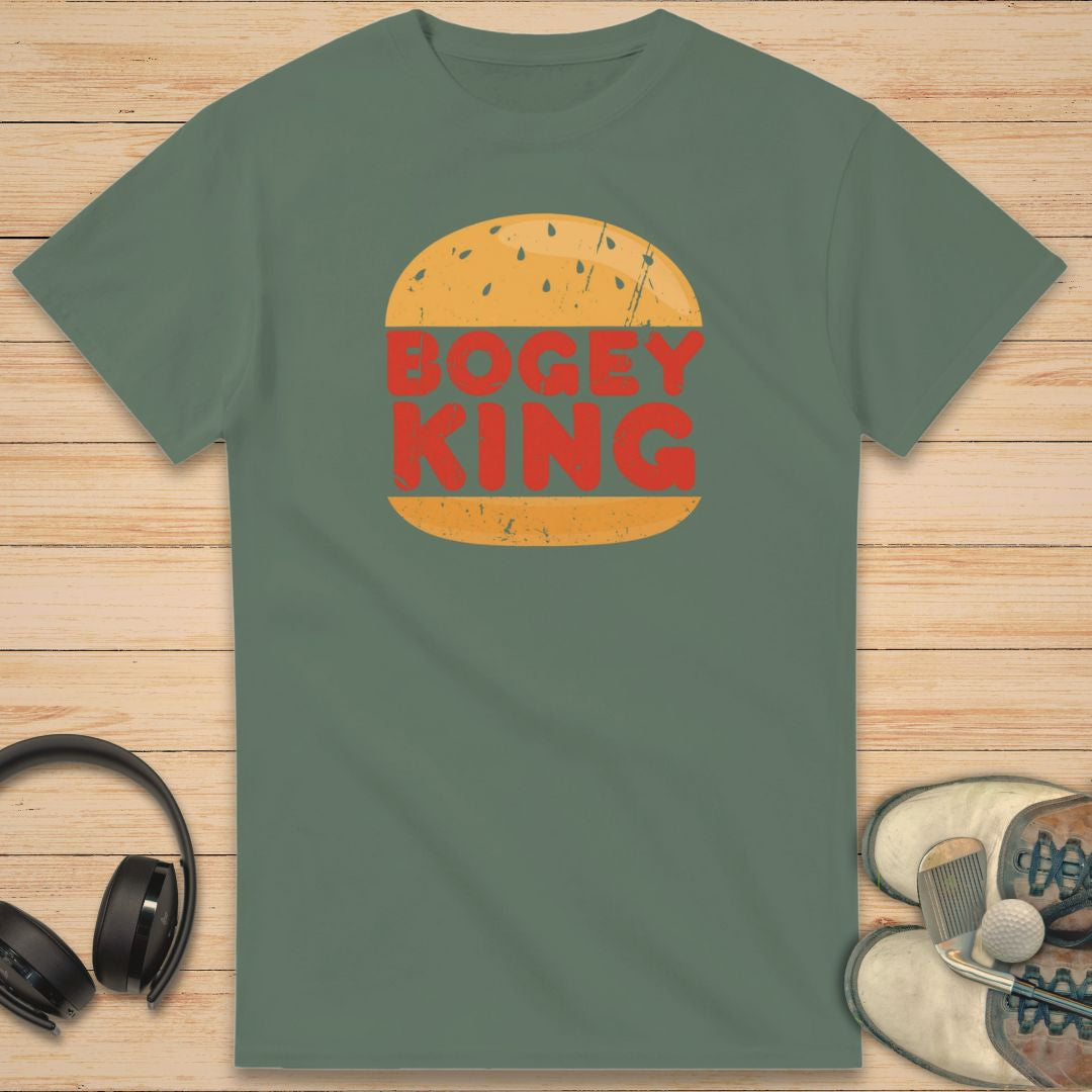 Bogey King T-Shirt in Military Green on a light wooden floor lying next to headphones and golf shoes.