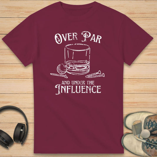 Over Par T-Shirt in Maroon on a light wooden floor lying next to headphones and golf shoes.