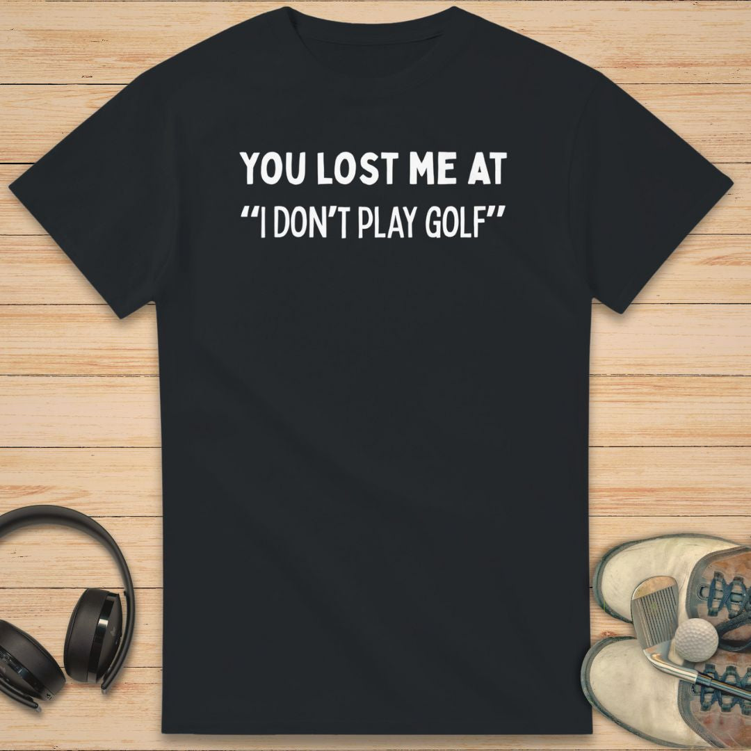 Lost Me T-Shirt in Black on a light wooden floor lying next to headphones and golf shoes.