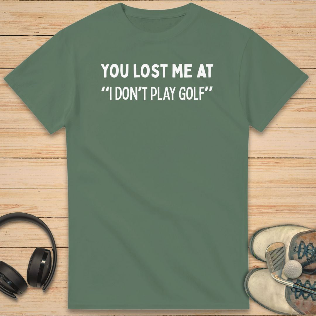 Lost Me T-Shirt in Military Green on a light wooden floor lying next to headphones and golf shoes.
