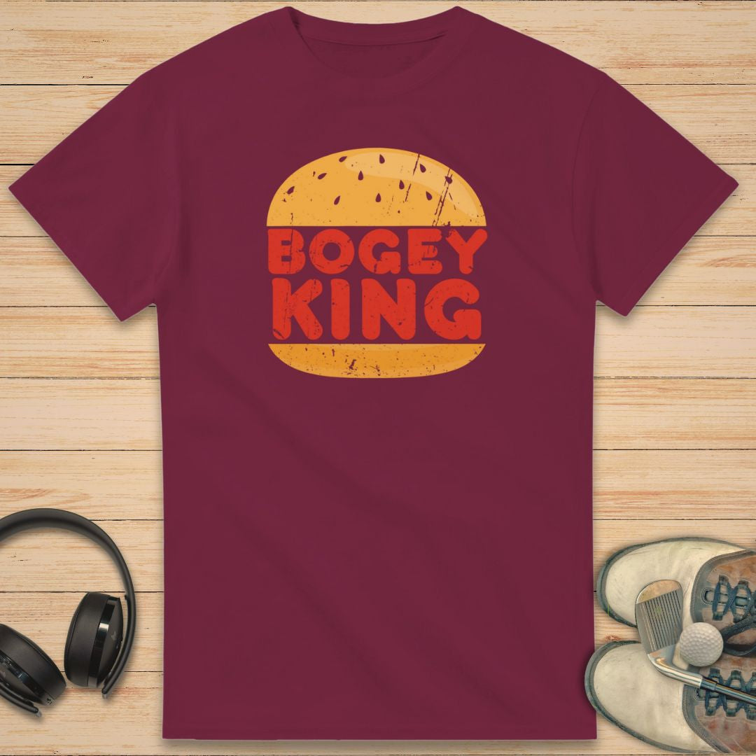 Bogey King T-Shirt in Maroon on a light wooden floor lying next to headphones and golf shoes.