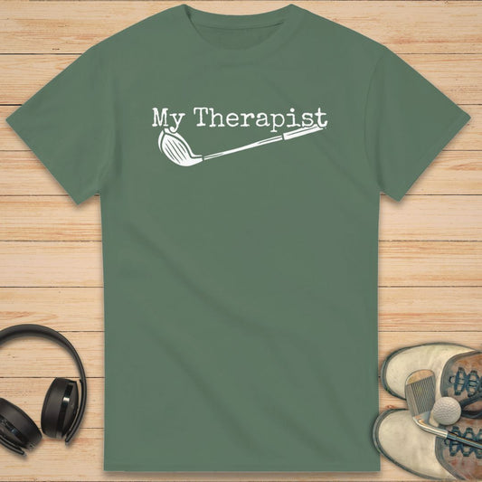 My Therapist T-Shirt in Military Green on a light wooden floor lying next to headphones and golf shoes.