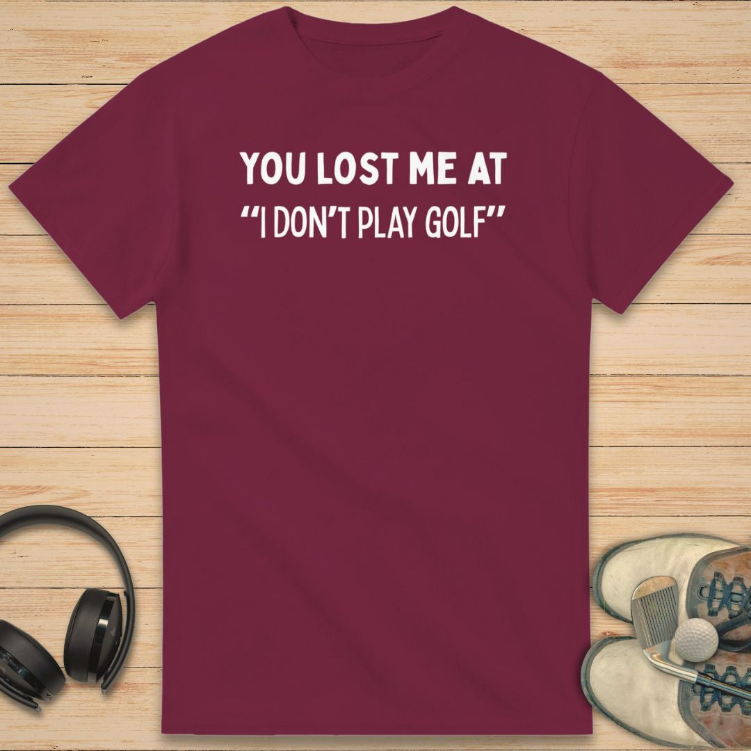 Lost Me T-Shirt in Maroon on a light wooden floor lying next to headphones and golf shoes.