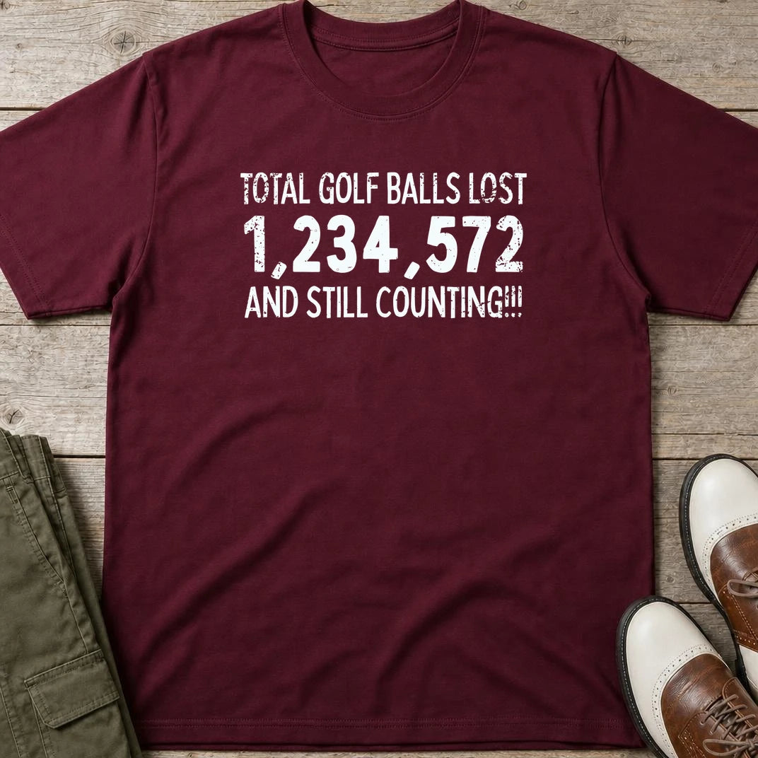 Golf Balls Lost 1,234,572 T-Shirt in Maroon lying on a plain floor. T-shirt is a unisex cut and 100 per cent cotton.
